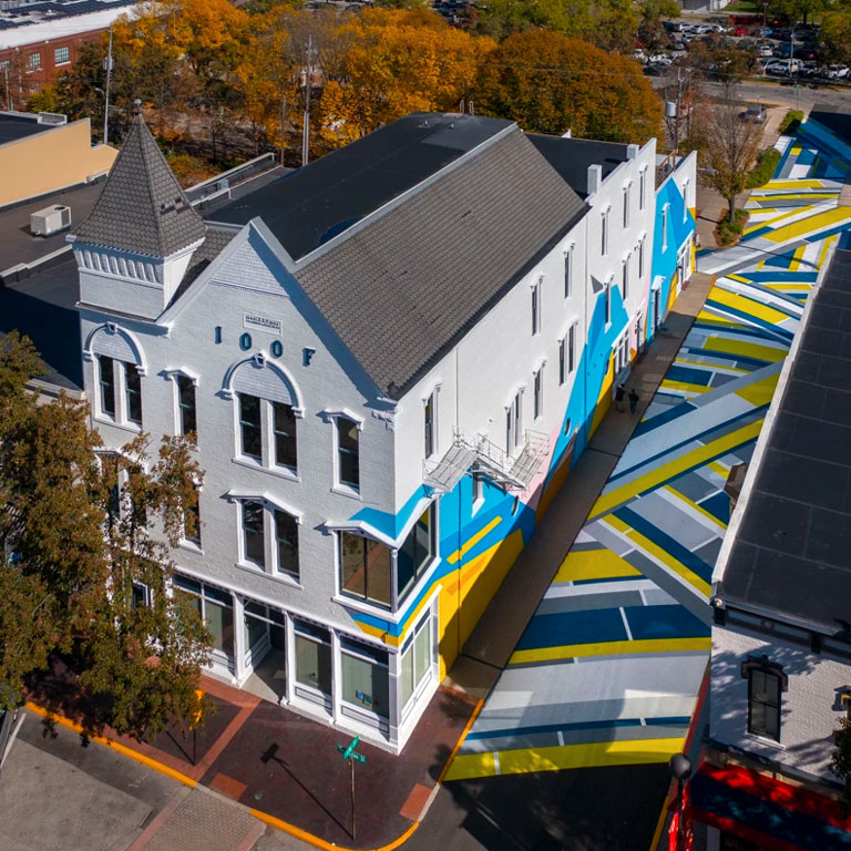 A geometric asphalt mural in bold shades of blue, yellow, white, and grey in an alley next to a large white building.