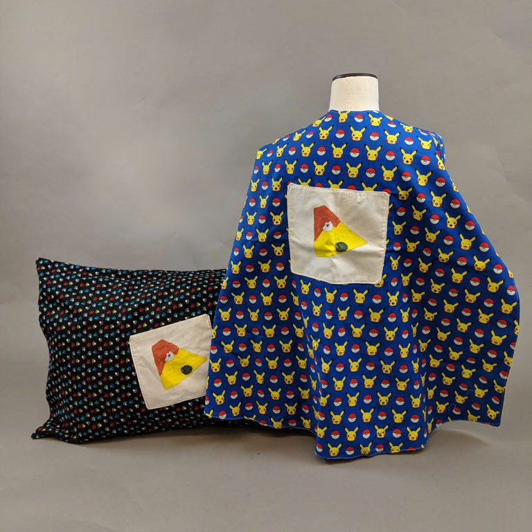 A children's cape and pillow case made out of Pokémon fabric, with a small patch on the back of the cape with a child's illustration of a Pokémon. 