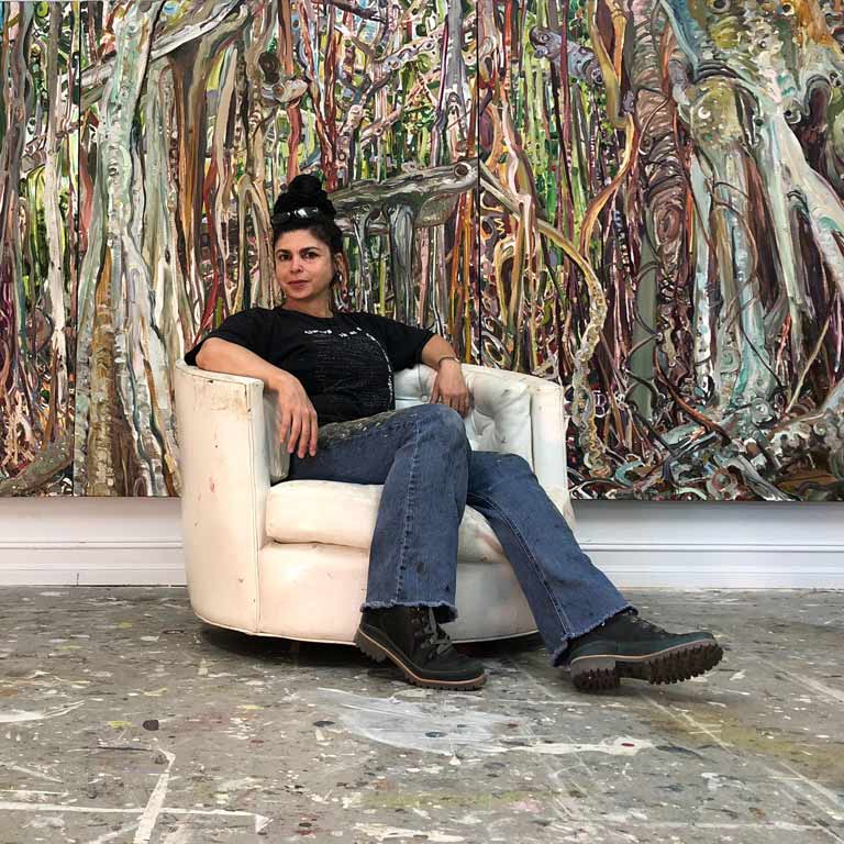 A portrait of Lilian Garcia-Roig sitting in a chair in front of their painting.