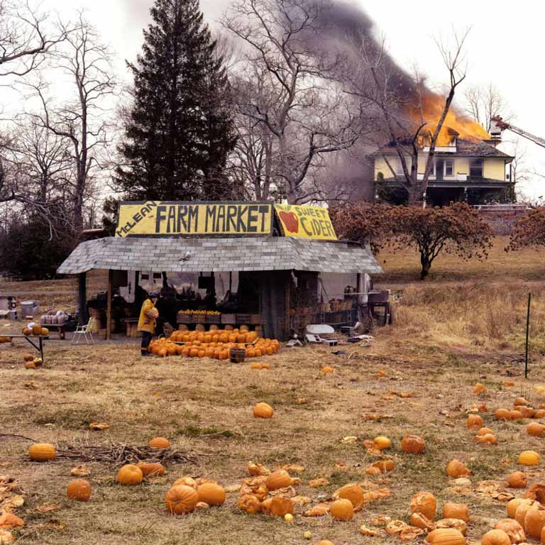 A farmer's market with pumpkin with a burning building in the background.