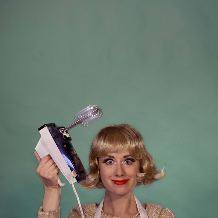 A blonde housewife holds up a hand mixer against a muted green background. 