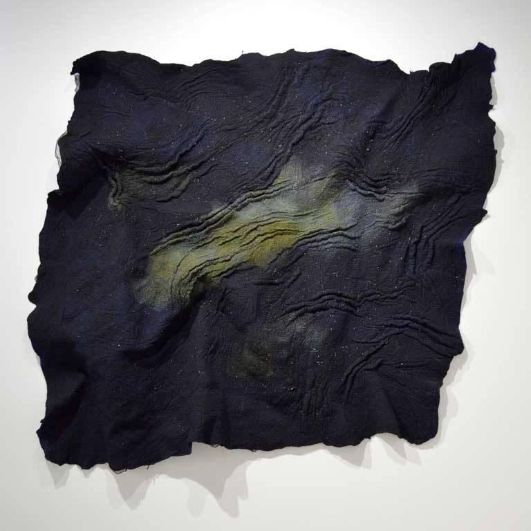 A navy blue fiber artwork hanging on a wall. It has various folds and waves set into it, culminating into a green center. 