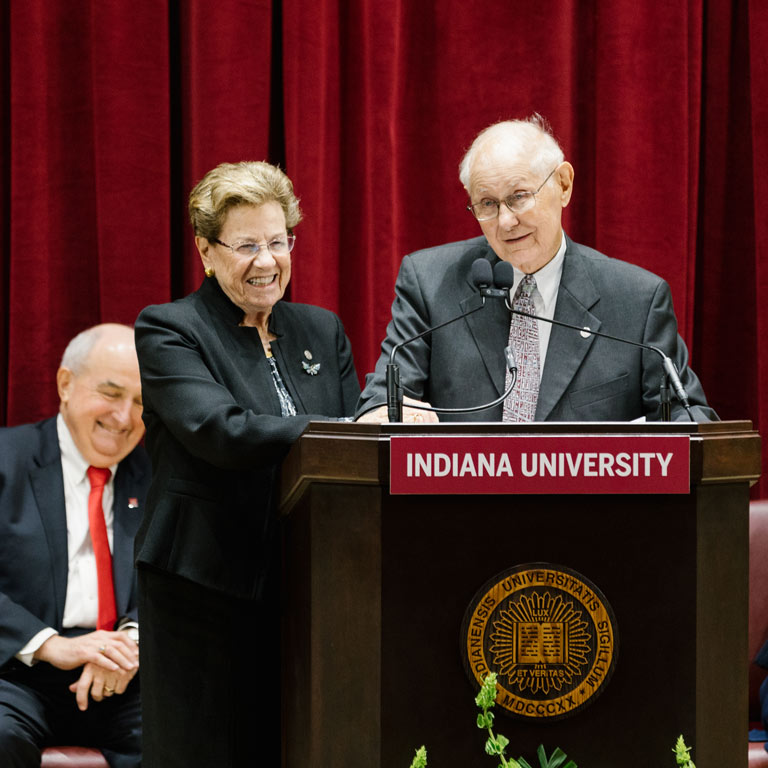Two older people smiling at a podium in front of a red velvet curtain.