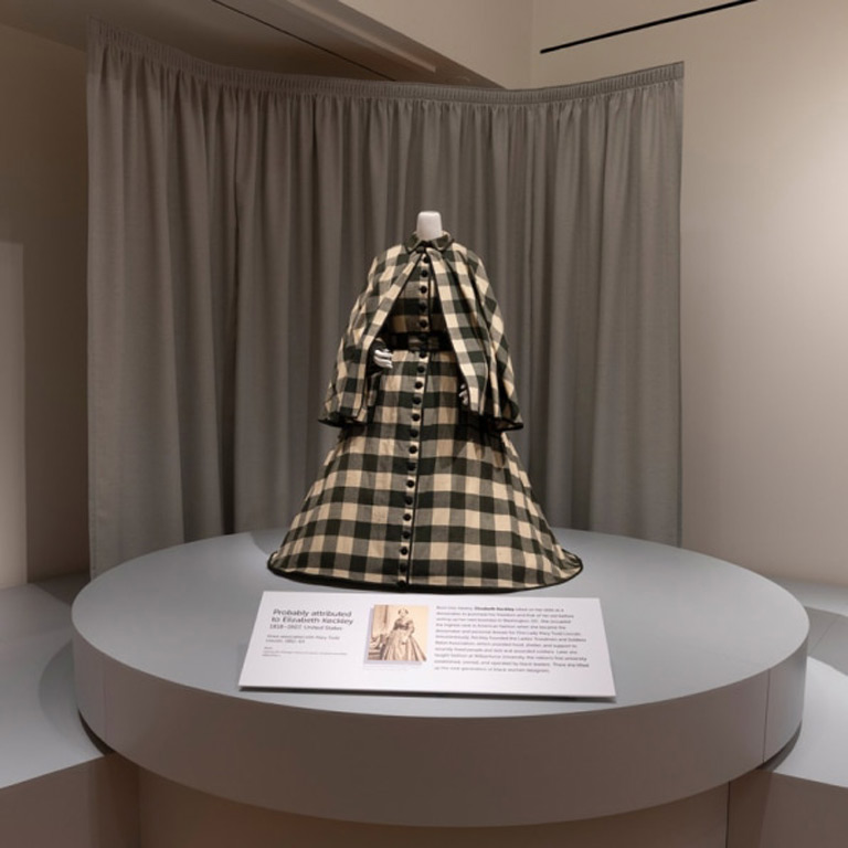 A black and white gingham dress and cape on a mannequin in a museum exhibition. 
