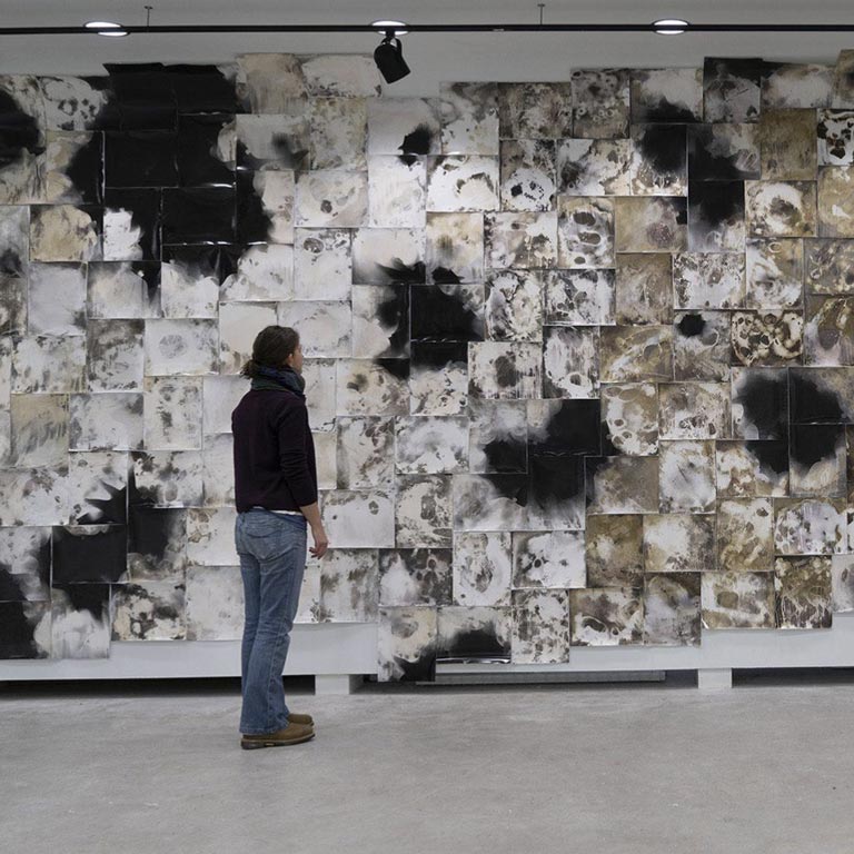 A person stands in front of a large black, white, and ochre artwork on the wall of a gallery.