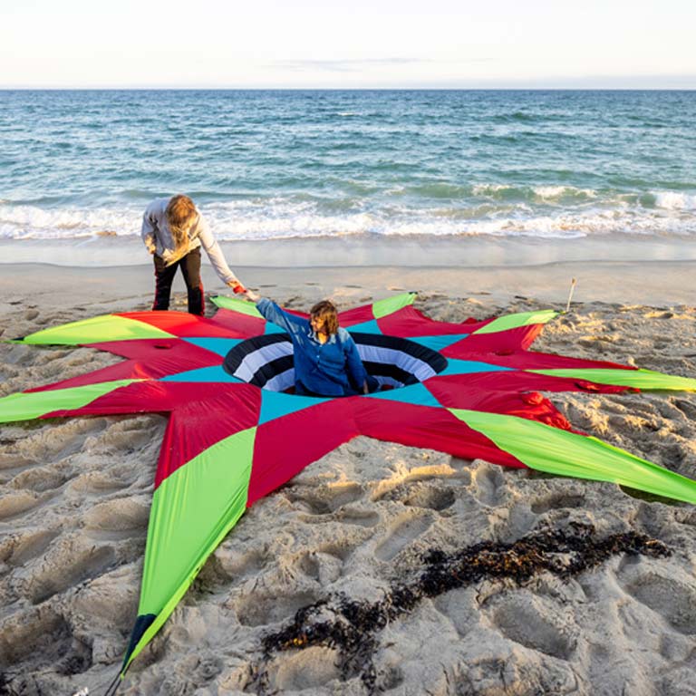 Two people install a large artwork on the beach. One of them stands in a hole they have dug with the center of the artwork down inside, the rest reaching outward. 