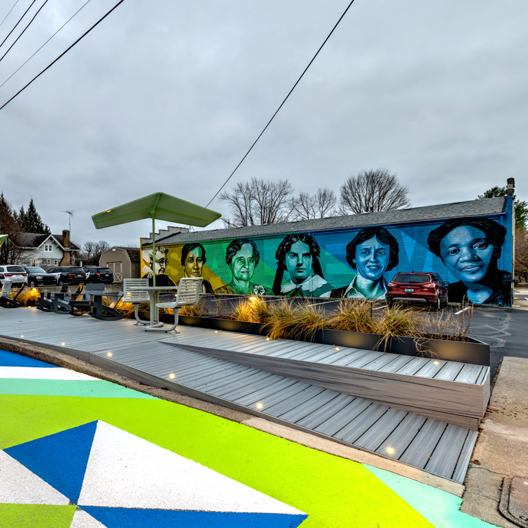 A large geometric mural painted in bright green, blue and white on asphalt, next to a mural of portraits of pioneering women from Salem, Indiana in similar colors.