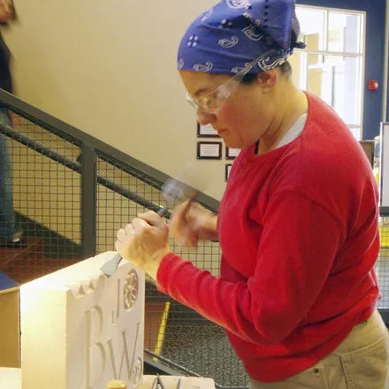 A person uses a chisel and hammer on a large piece of limestone. They are wearing protective glasses, a blue bandana over their hair, and a red sweatshirt. 