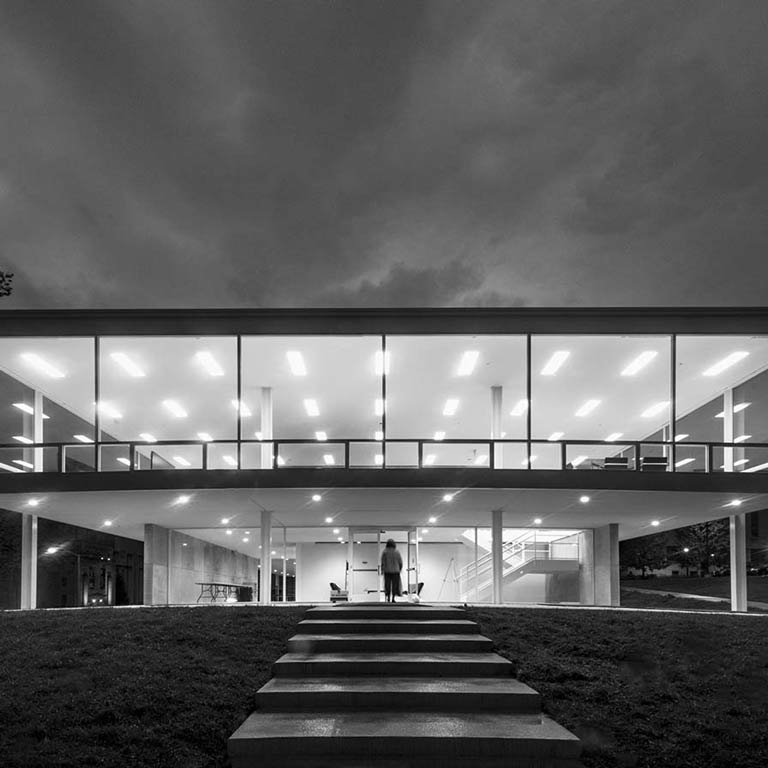 A black and white photo the Mies van der Rohe building lit up at night with a person walking up the main staircase.
