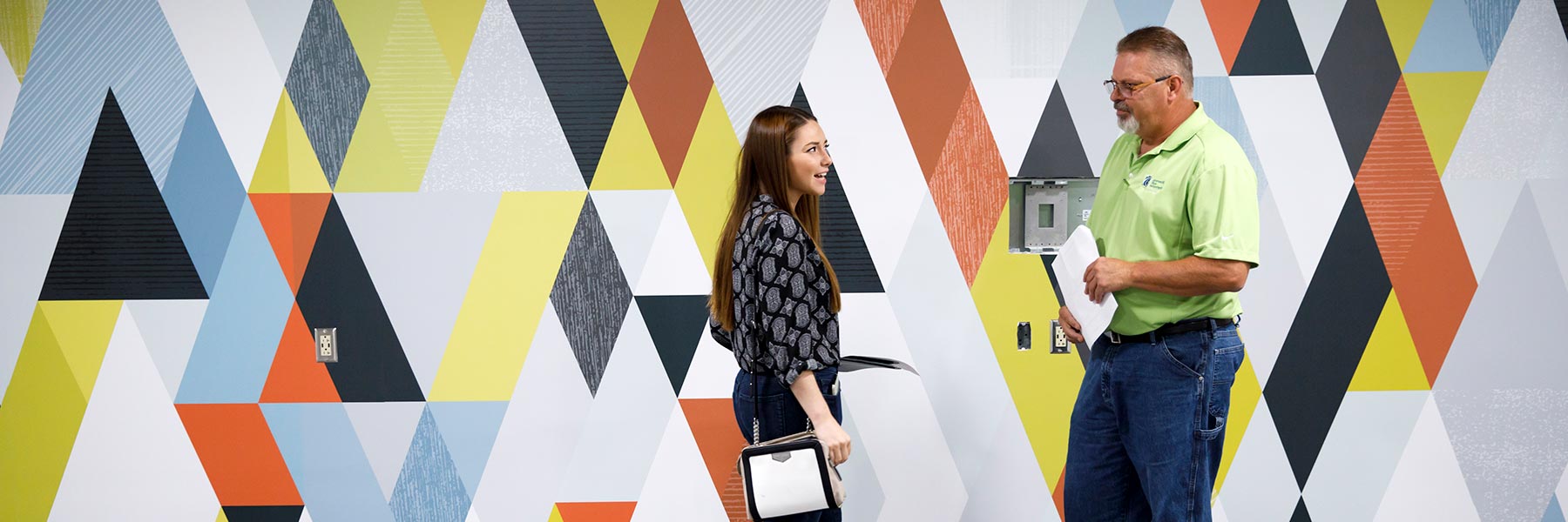 Two people stand in front of a patterned wall.